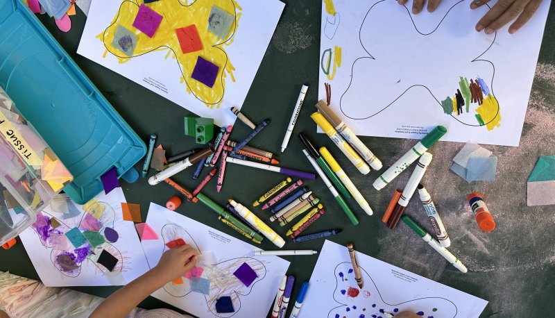 Close-up of children's hands working on an art project. The hands are coloring butterfly templates with markers and crayons, and adding tissue paper to the designs. The materials are scattered around, showcasing the creative process.