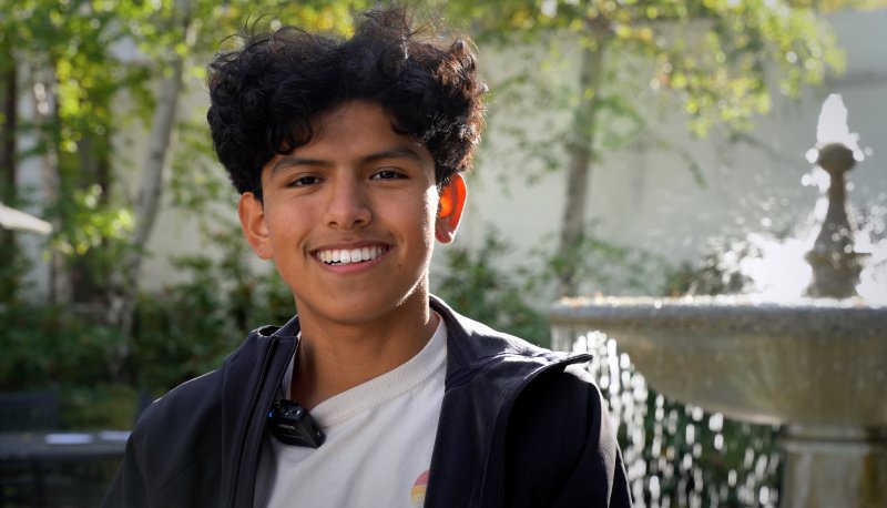 Mitchell Larrieta Angeles smiles at the camera, with a fountain behind him