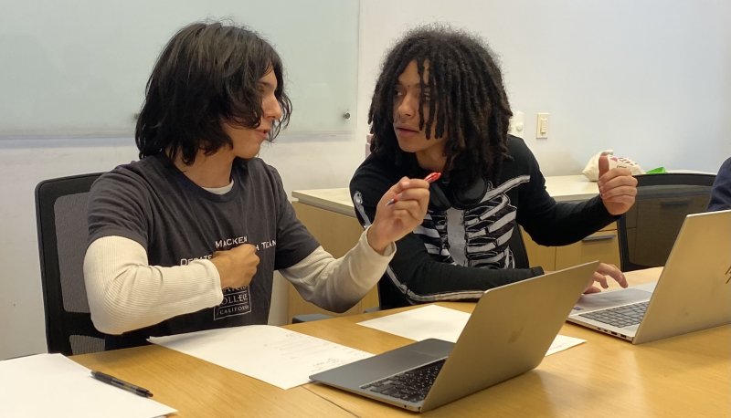 A young light brown man gestures with his hands on the left while a young Black adult looks intently at him while also gesturing. They both have laptops open in front.