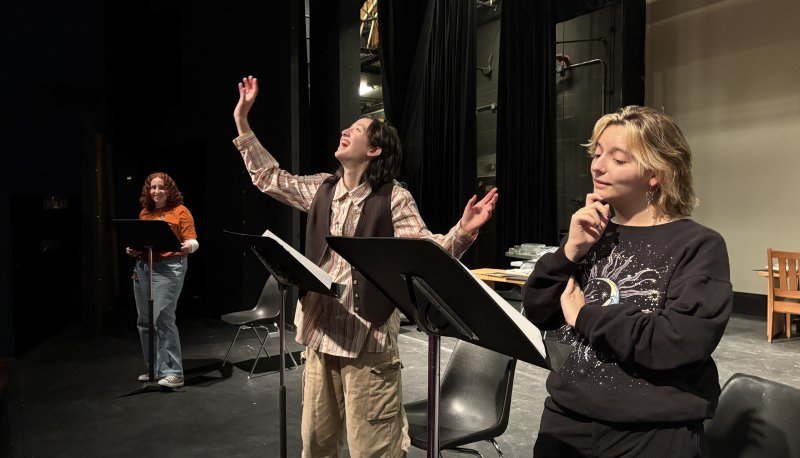 Three students rehearse a staged reading for TAKE.UP.SPACE. with music stands holding their scripts, and an actor dramatically looking up with arms outstretched.