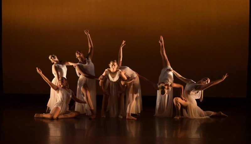 a group of modern dancers in flowing white dresses pose with a burnt sienna background.
