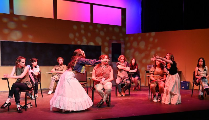 Actors perform in a play where students are seated in a classroom and two female characters are dancing in white long skirts together.