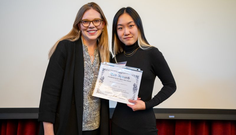 Student holds up Best Research certificate on stage with Dean Lauren MacDonald