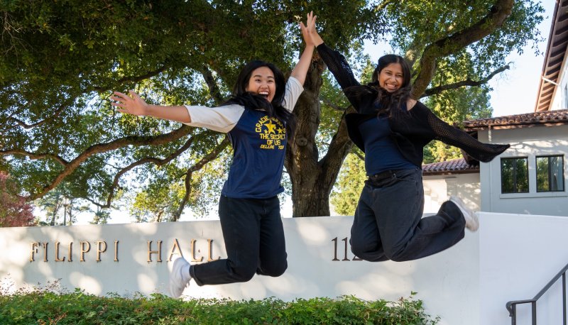 Two students jumping in front of Filippi Academic Hall in fall 2025