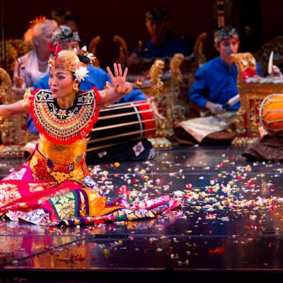 Balinese Dancer in colorful outfit, palms outreached, sitting on the floor with confetti. Instruments being played in the background
