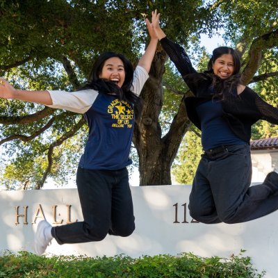 Two students jumping in front of Filippi Academic Hall in fall 2025