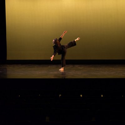 A dancer in black bends forward at the waist and lifts their leg into an attitude position, against a yellow background