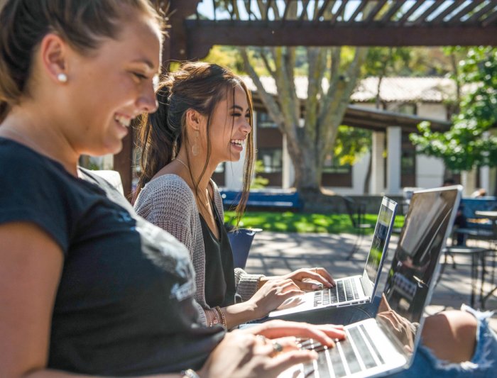 Two students laughing while on their laptops outside