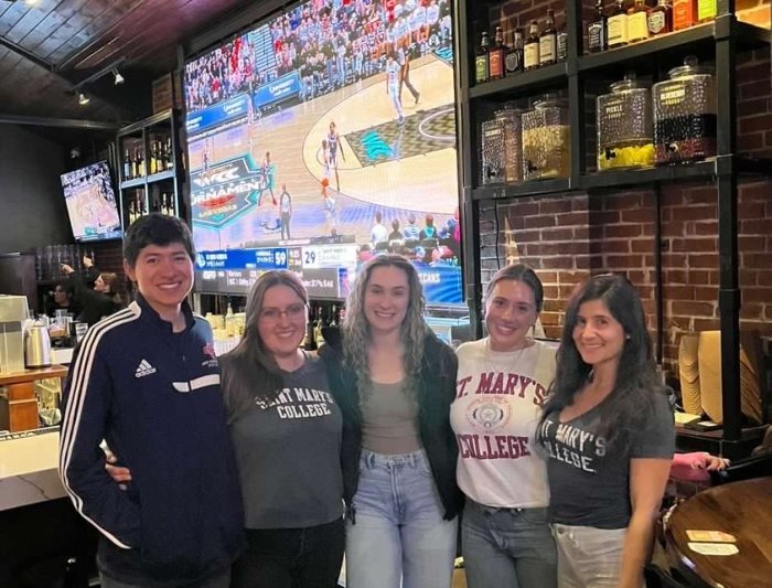 Five people wearing Saint Mary's gear in front a big screen showing basketball