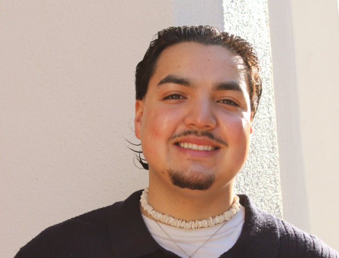 Smiling person with short dark hair and facial hair, wearing a dark sweater and a shell necklace, standing against a sunlit textured wall.