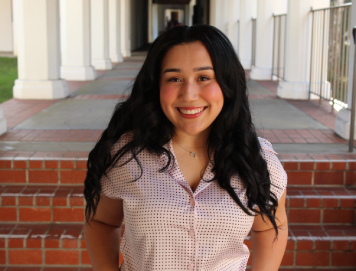 A smiling woman with long black hair stands outside in a hallway with white columns. She is wearing a pink polka-dot blouse, conveying a cheerful mood.