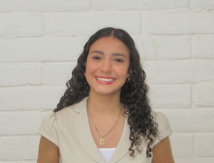 Smiling student staff member with curly dark hair wearing a light-colored blouse, standing against a white brick wall.