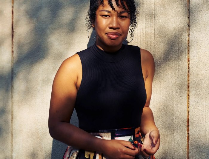 Headshot of woman standing infront of a wooden fence, in a black top and printed skirt