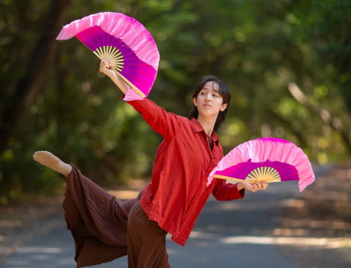 Anya tang poses outdoors with trees in the background and Japanese fans in his hands