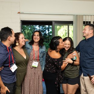 Six people embrace and laugh inside a townhouse living room