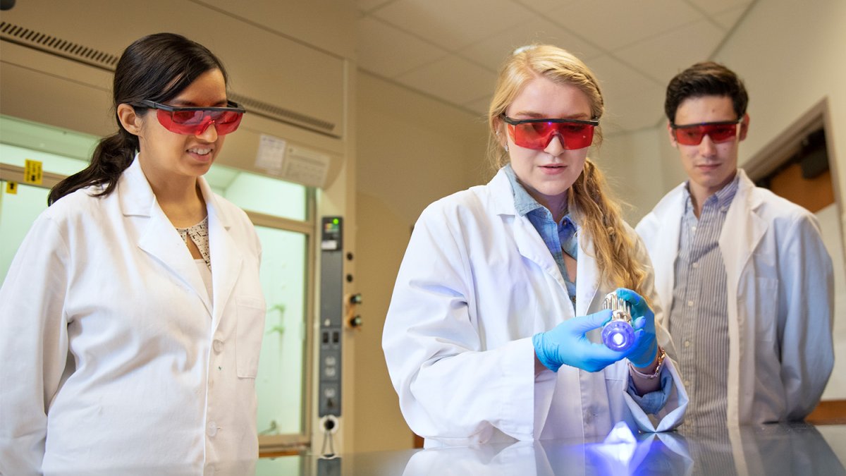 students in lab coats and protective glasses work on an experiment