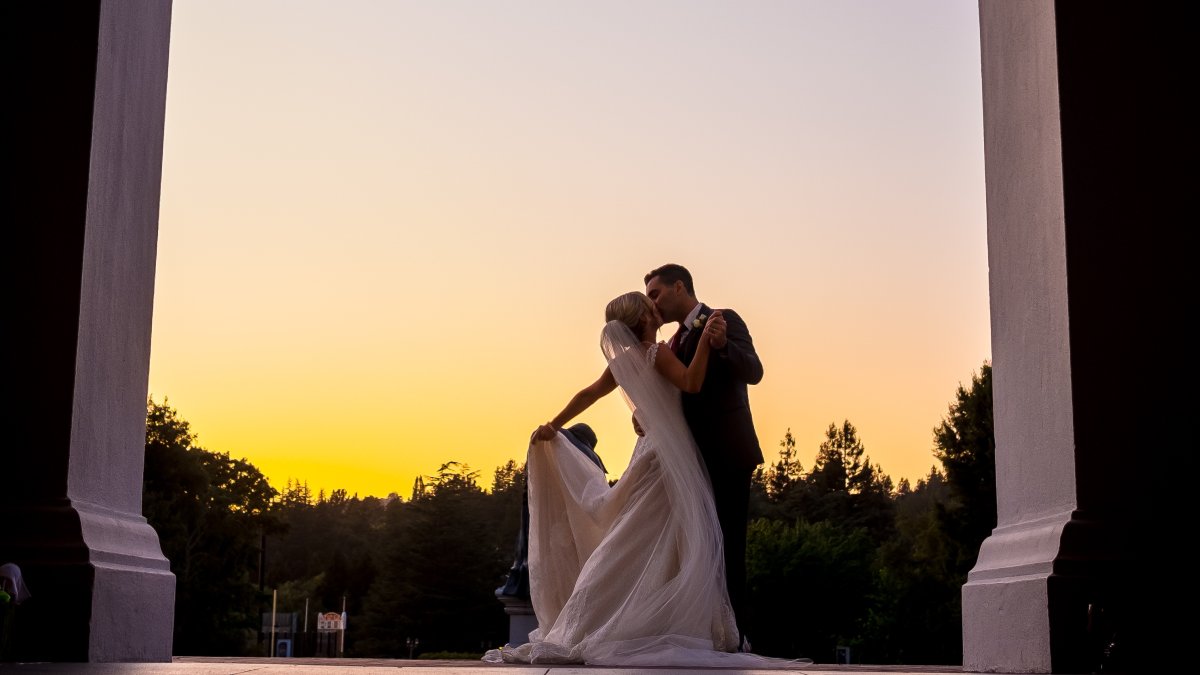 Image of a wedding couple kissing. An archway is at the forefront of the image with the couple behind, and they are silhoutted by the setting sun.